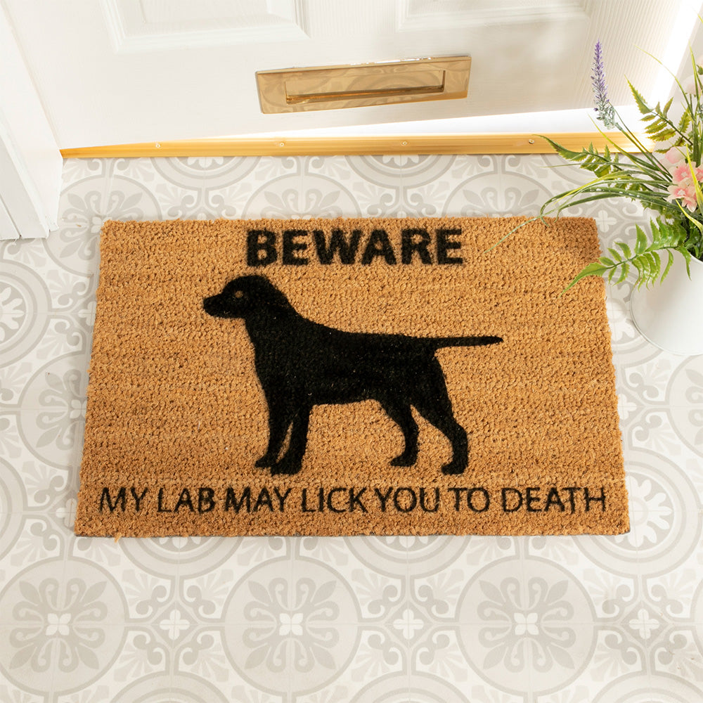 Coir doormat featuring the silhouette of a Black Labrador and the words 'Beware my lab may lick you to death'. The mat is in front of a door with a plant next to it.