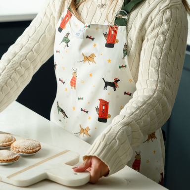 A person wearing a white apron with dark green straps. The apron features a printed design with different dog breeds and post-boxes at Christmas. She is in the kitchen holding a tray of mince pies.