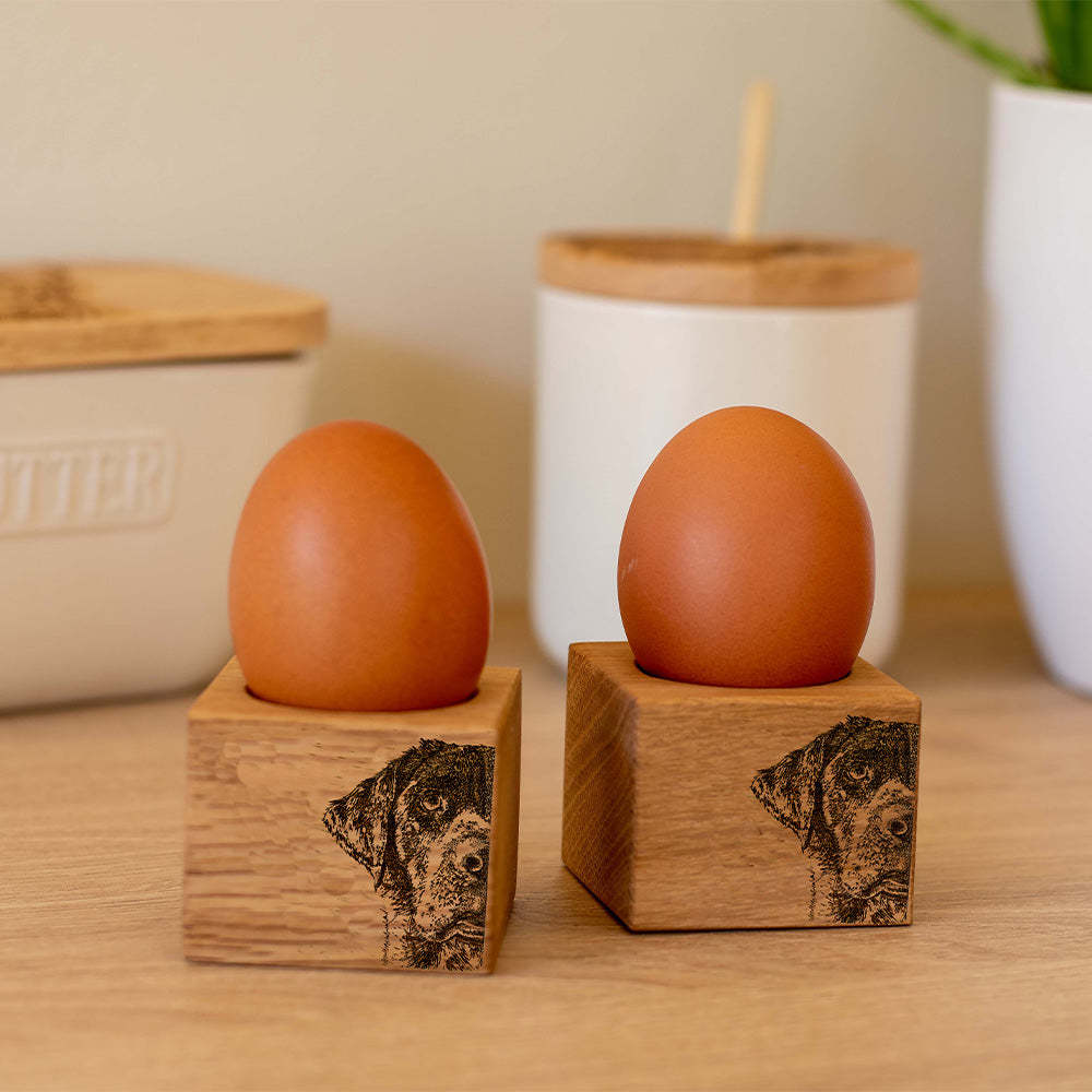 Two oak egg cups with an engraving of half a Labrador's face. They are on a kitchen counter with an egg in each.