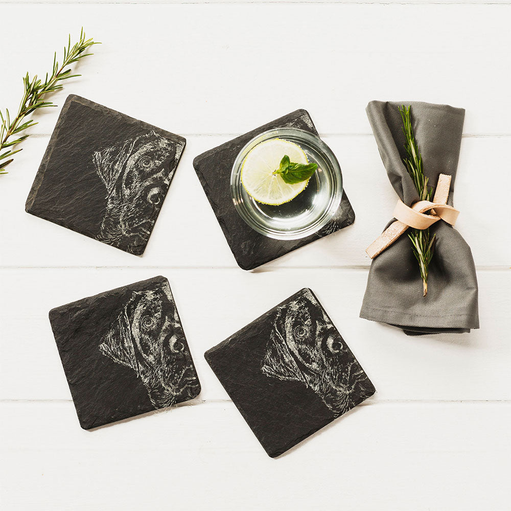 Four slate coasters each featuring an engraving of a Labrador's head, are seen on a tabletop with glass and napkin next to them.