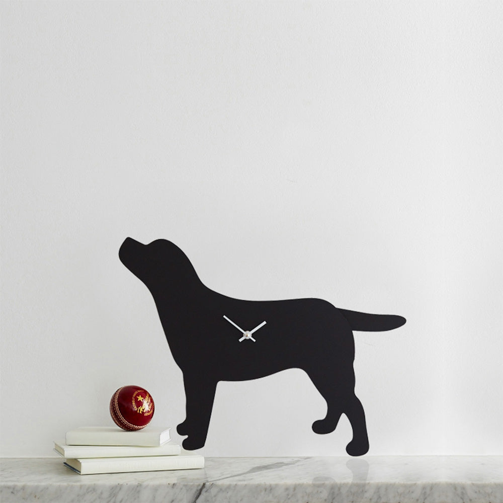 A photograph of a clock that has the silhouette of a Black Labrador.  It has two white clock hands in the middle. It has been placed next to three books and a ball.