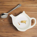 A white china teabag tidy in the shape of a teapot, with Labrador puppy and rope toy design. There is a spoon next to it on a wooden background.