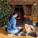A woman wearing a bright blue jumper with white woven pattern featuring leaping puppies and snowflakes is sitting with a golden labrador in front of a Christmassy fireplace.