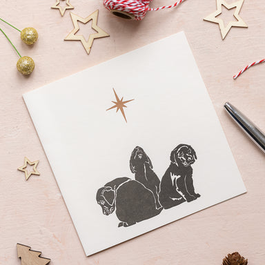 A white card with 3 black puppies and gold star sits on a Christmas table scattered with small gold star decorations