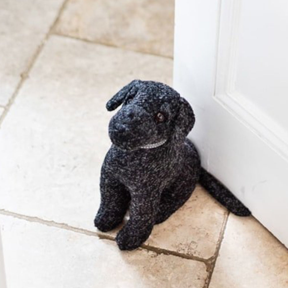 A black fabric door stop in the shape of a Labrador. The door stop is holding open a white door.