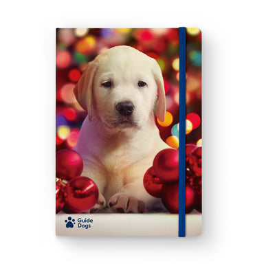 A close up of a Guide Dogs Christmas notebook. The front of the notebook has an image of a yellow labrador puppy surrounded by red baubles and lights in the background. The Guide Dogs logo is in the bottom left corner and blue elastic holds the pages closed.