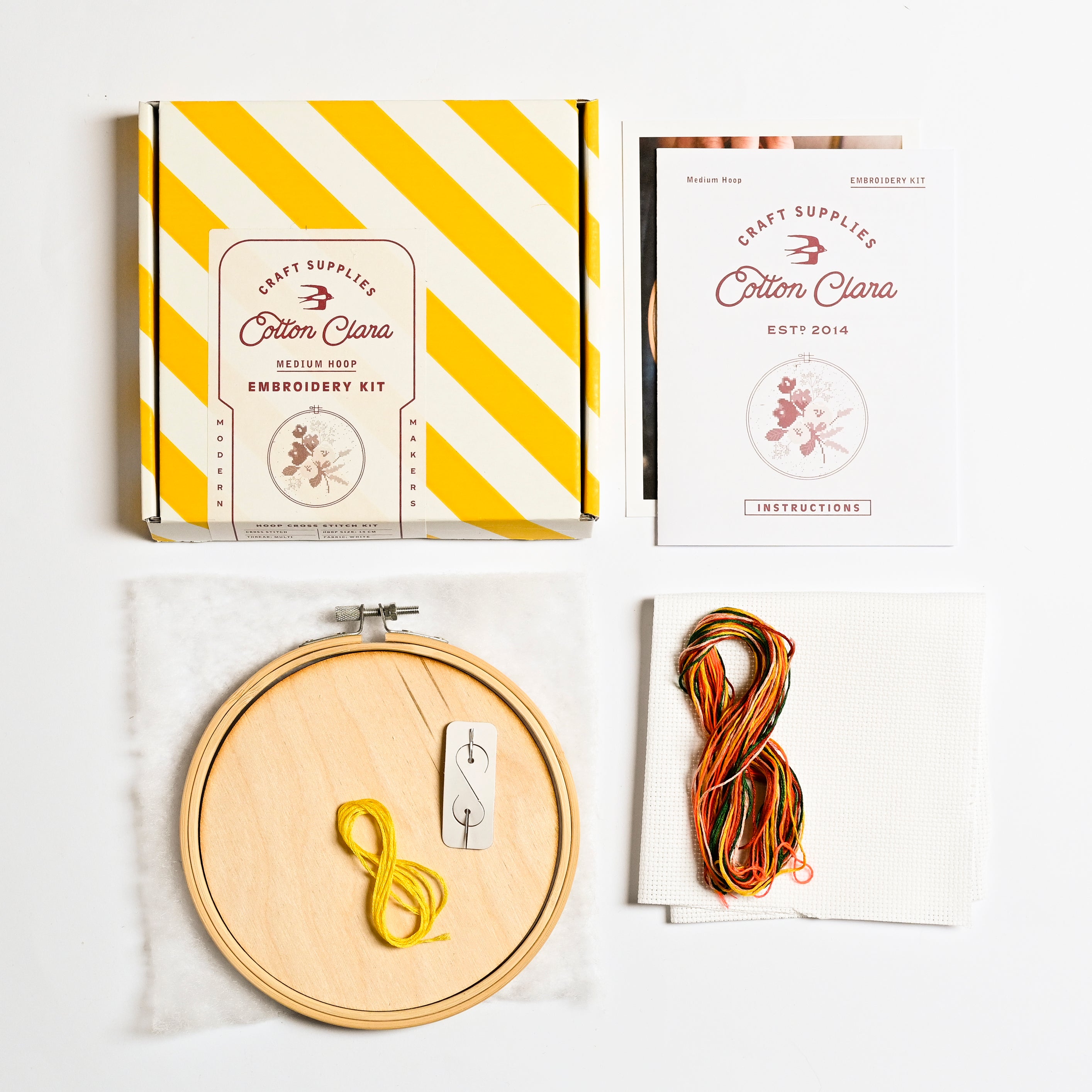 The embroidery kit box and it's contents are displayed. The box has yellow and white stripes, with instructions on white card with orange text. A embroidery hoop has a loop of yellow string whilst a collection of yellow and orange strings are in a loop together.