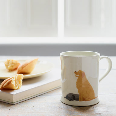 A white ceramic mug with two dogs on. A happy Golden Retriever sits next to a sleeping Black Labrador.