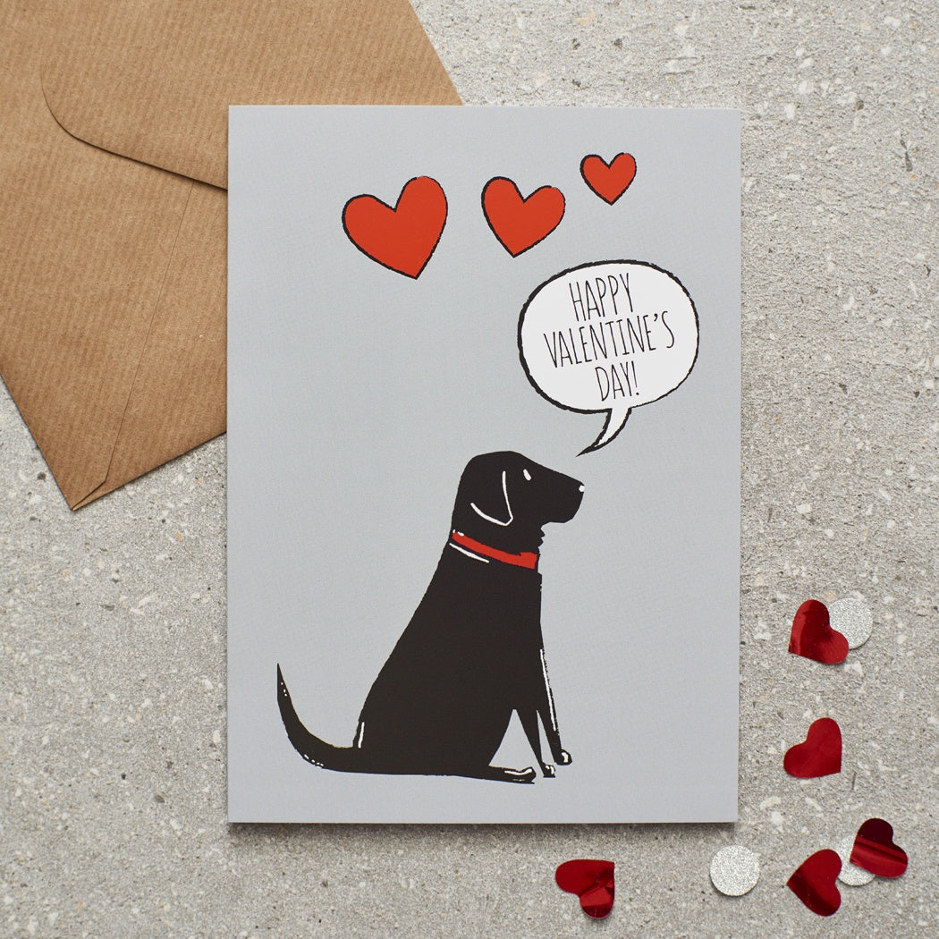 A greetings card with a Black Labrador with a red collar and a speech bubble saying Happy Valentines Day and three red hearts. Behind the card is a brown paper envelope on a textured background.