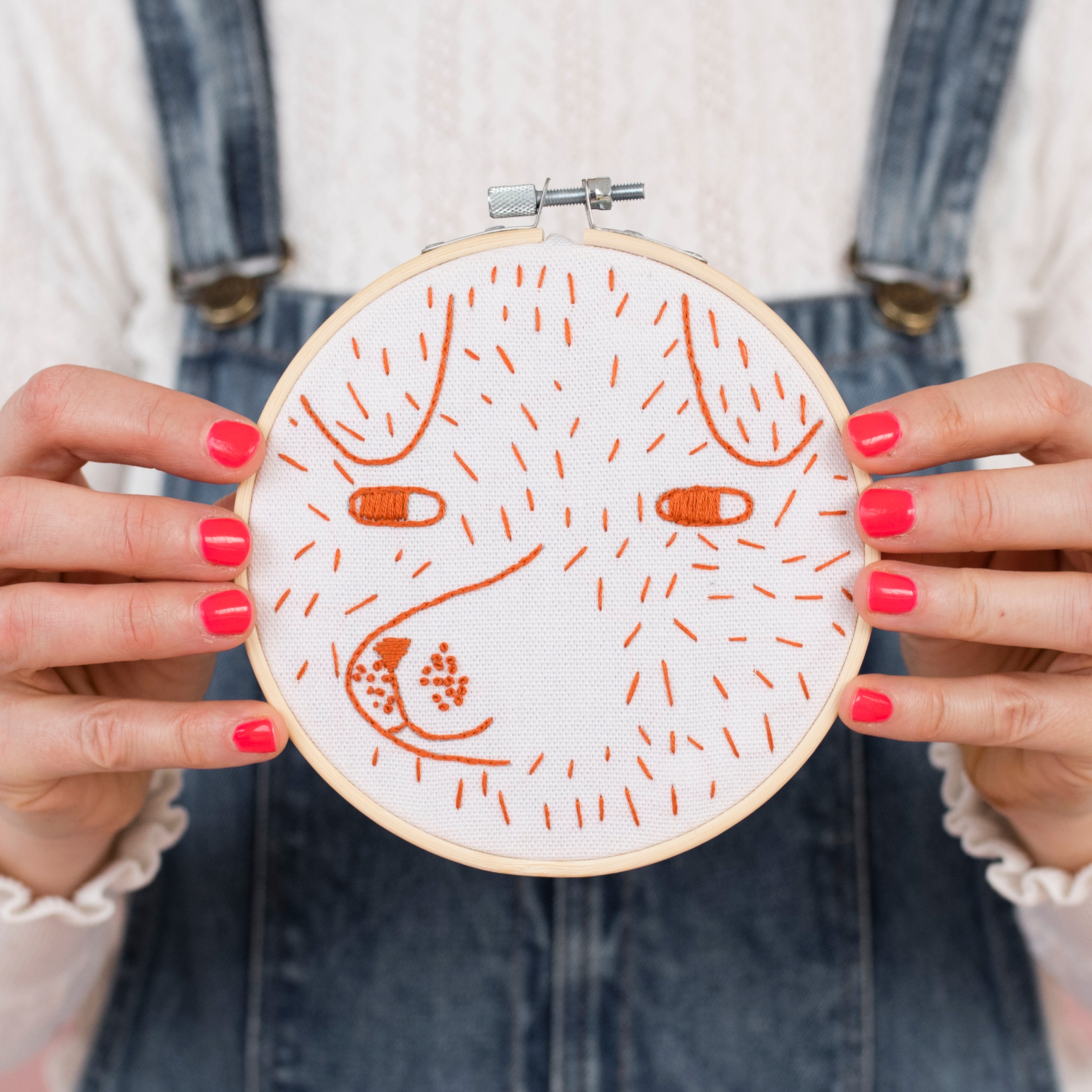 A lady in a white lace top and blue denim dungarees with painted orange nails holds an embroidery hoop in front of her. The hoop shows a pattern of a dog in orange string stitched onto the material.