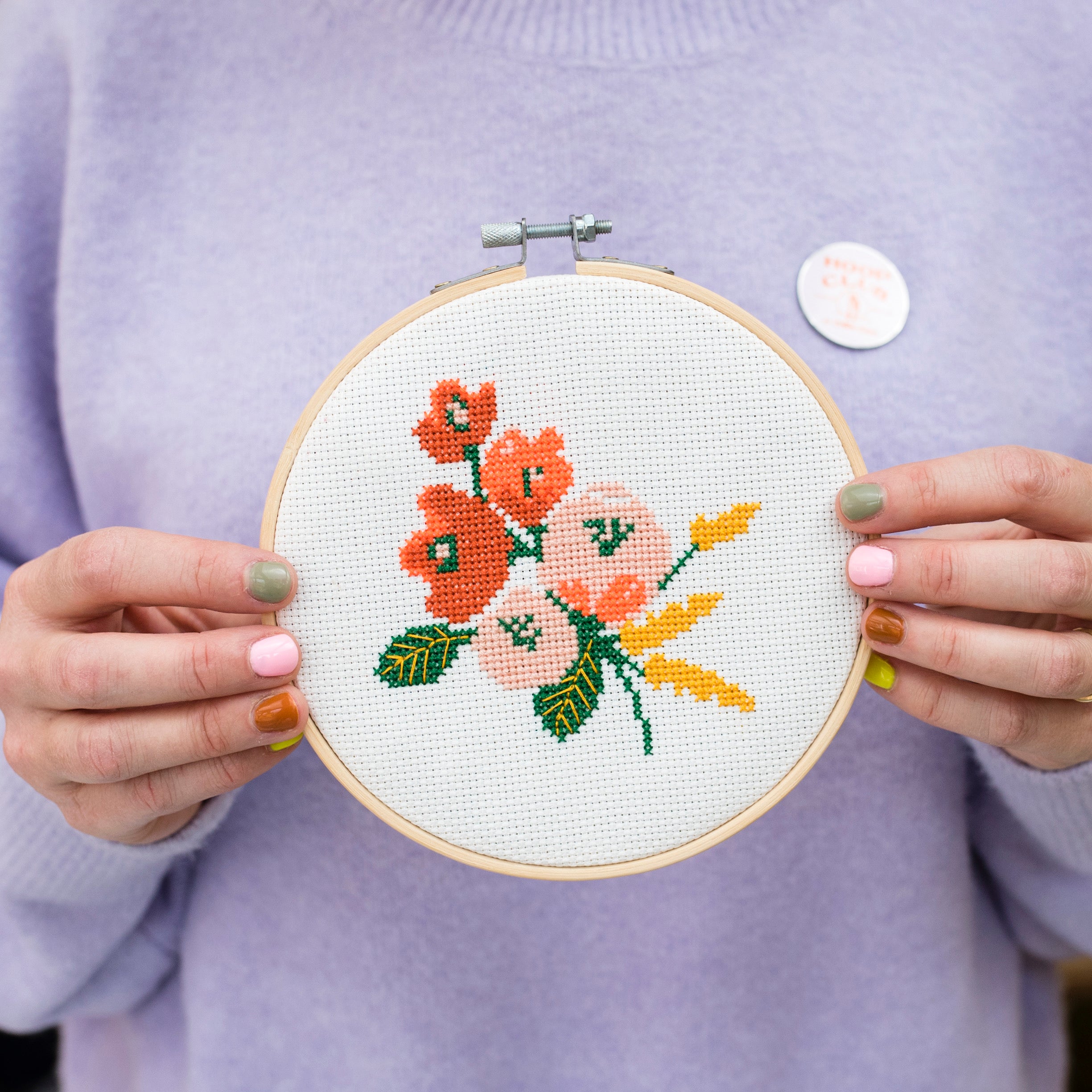 A close up of a lady in a purple jumper with painted nails holding an embroidery hoop with a floral pattern.
