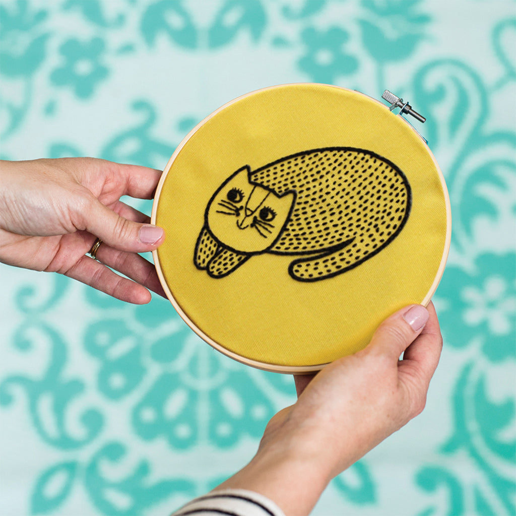 A lady in a black and white stripey top holds an embroidery hoop with a black cat deisign stitched onto yellow material. In the background is a blue floral pattern on a white base.