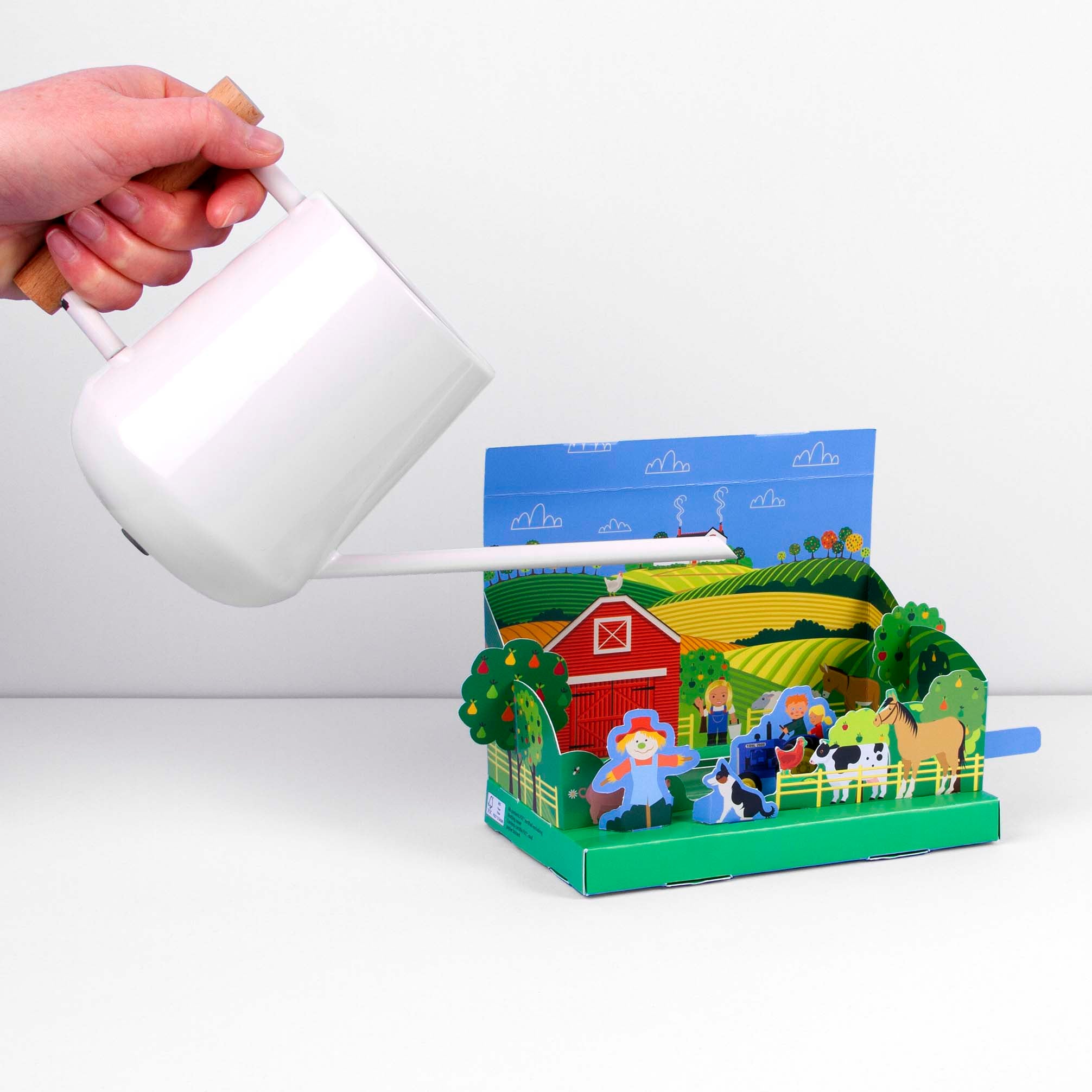 A hand holding a white watering can is shown watering the children's mini farmyard garden