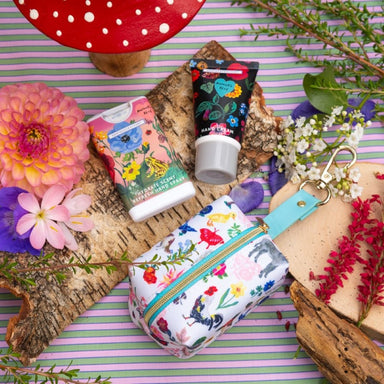 A floral hand spray and hand cream next to a treats bag printed in a bright floral design, surrounded by flowers and leaves