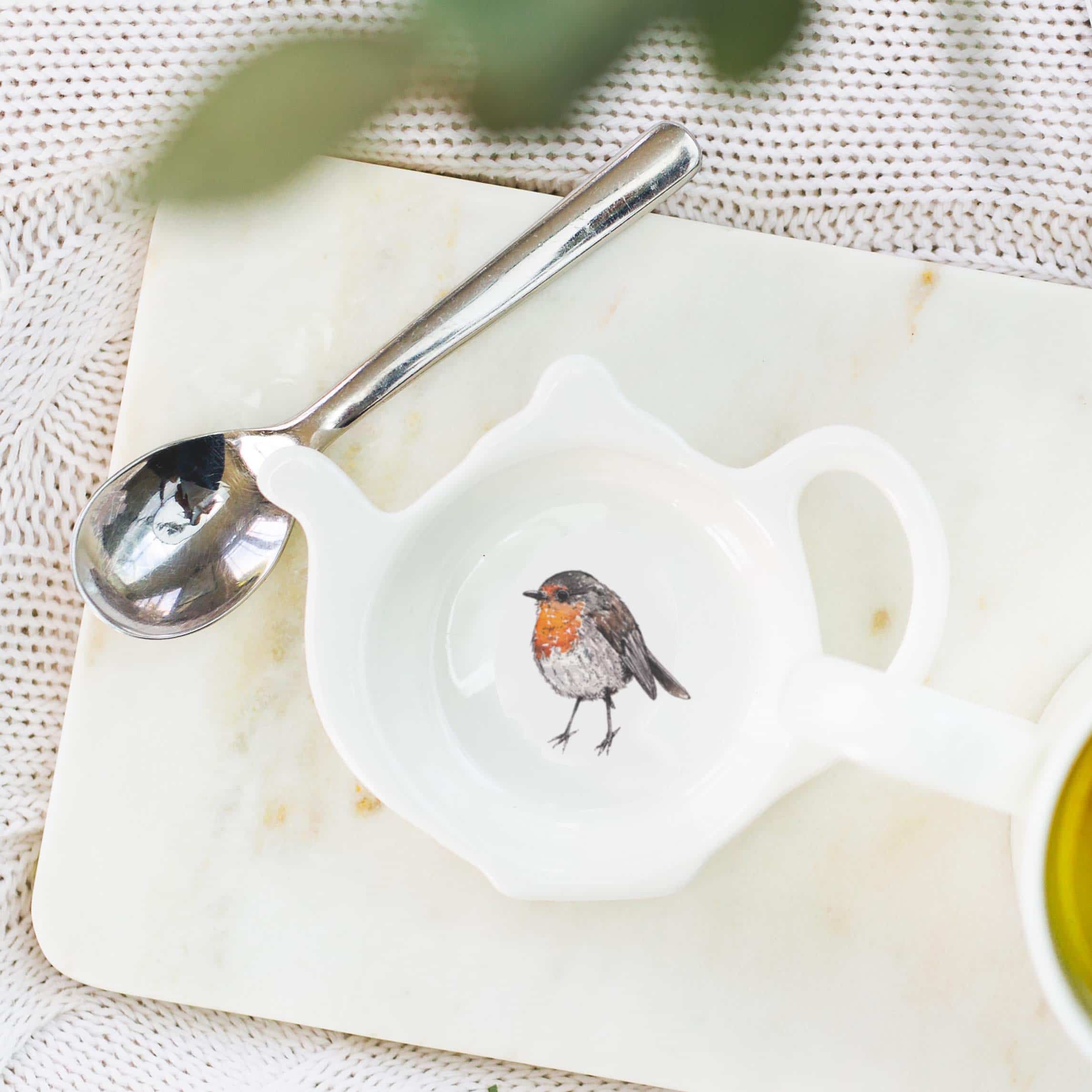 White ceramic tea bag tidy with robin design sitting on a white tile with teaspoon next to it