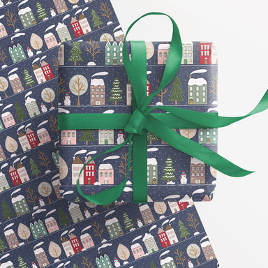 A piece of wrapping paper appears underneath an already wrapped present with green ribbon. The wrap is blue with illustrations of snowy houses and trees.