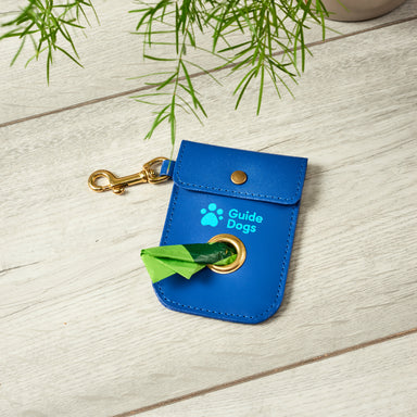 A blue leather poo bag dispenser that has a brass clip to attach to a lead or bag is lay on wooden floor. The Guide Dogs logo is on the pouch, above the hole for easy removal of poo bags.
