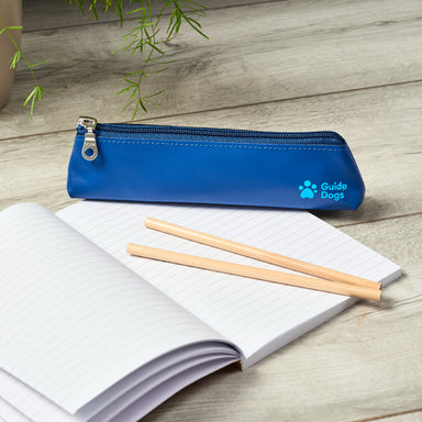 A blue leather pencil case rests on top of an open lined notebook with two pencils. The zipper on the case is silver and the Guide Dogs logo is printed on the bottom right hand side of the pencil case.
