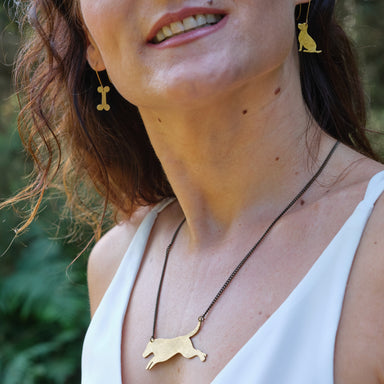 A lady with brown wavy hair wears brass jewellry. In her right ear she is wearing the brass bone earings, in the left ear a dog shaped earring. She is also wearing the leaping dog brass necklace.