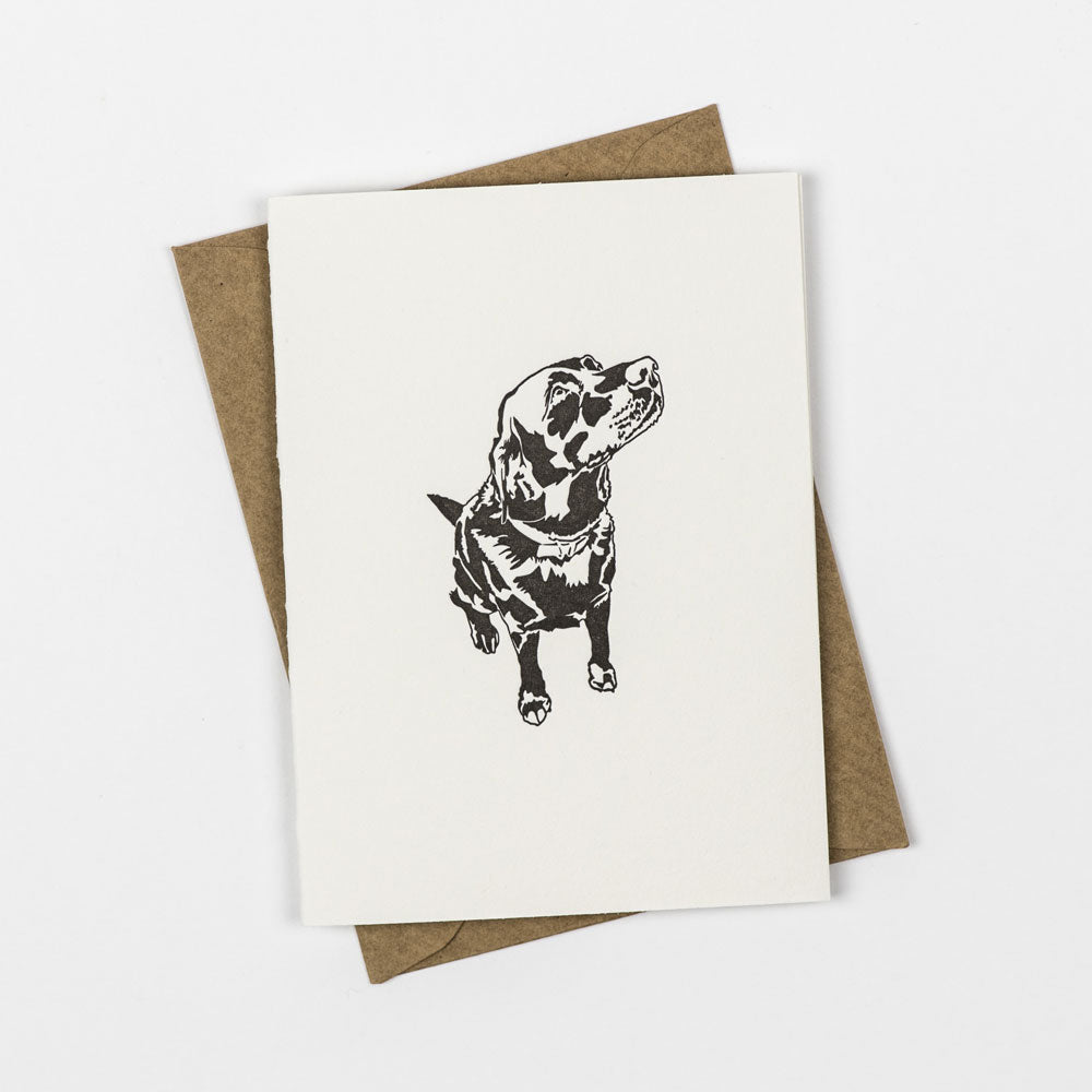 A white greetings card with a labrador design in the middle is above a brown kraft envelope on a white background.