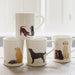 A collection of four white ceramic mugs. A mug with one Chocolate and one Black Labrador puppy playing is stacked onto two mugs. On the left, a white mug with a sleeping Black Labrador and a sitting Golden Retriever. On the right, a standing Black Labrador. To the right of the stack is a mug with a Yellow Labrador and two Labrador puppies, one Chocolate, one Black.