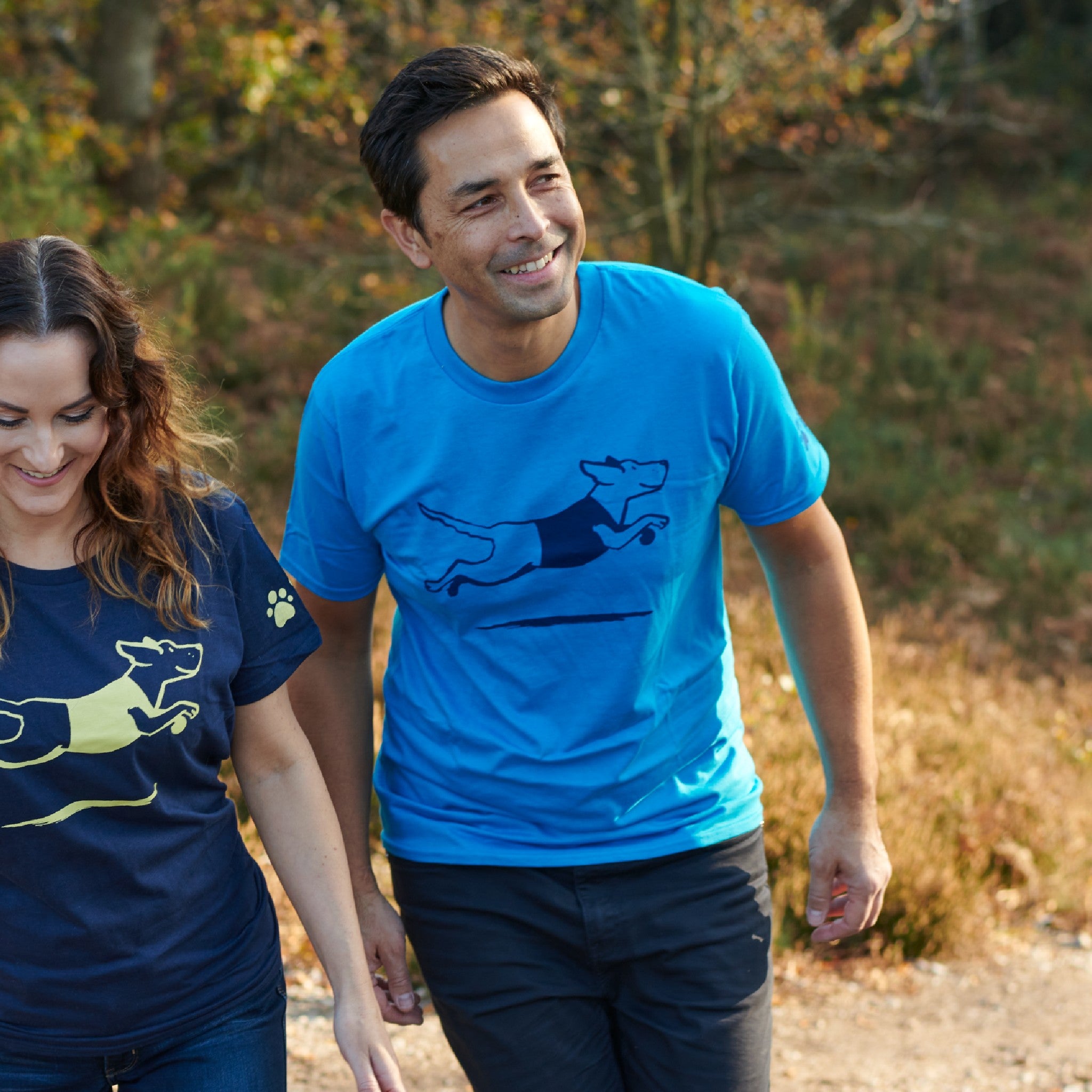 A man is outside wearing a light blue tshirt with a dark blue leaping dog design on the front. He is with a woman who is wearing a matching tshirt in navy and yellow.