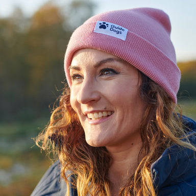 A woman with brown curly hair is wearing a light pink beany hat with the Guide Dogs logo on.