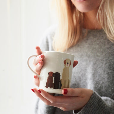 A lady with blonde hair and red nails is holding a white ceramic mug decorated with an illustration of three dogs, a Chocolate Labrador puppy, a Black Labrador puppy and a Yellow Labrador adult all sat down.