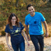A lifestyle image of a couple wearing Guide Dogs tshirts. The woman, on the left is wearing the navy and the man, on the right is wearing the blue. They are both walking through woodland.
