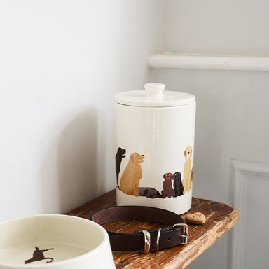 A white ceramic treat jar sits on a shelf with a black leather collar and a white ceramic dog bowl. The treats jar is decorated with illustrations of a Golden Retriever, a sleeping Black Labrador, two Labrador puppies, one Chocolate and One Black and a Yellow Labrador.