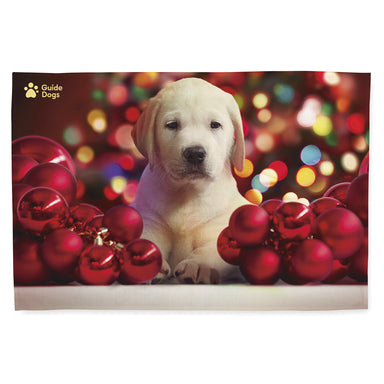 The Guide Dogs Christmas tea towel with the Yellow Labrador Puppy surrounded by red baubles and festive lights design. The Guide Dogs logo is in the top left hand side.