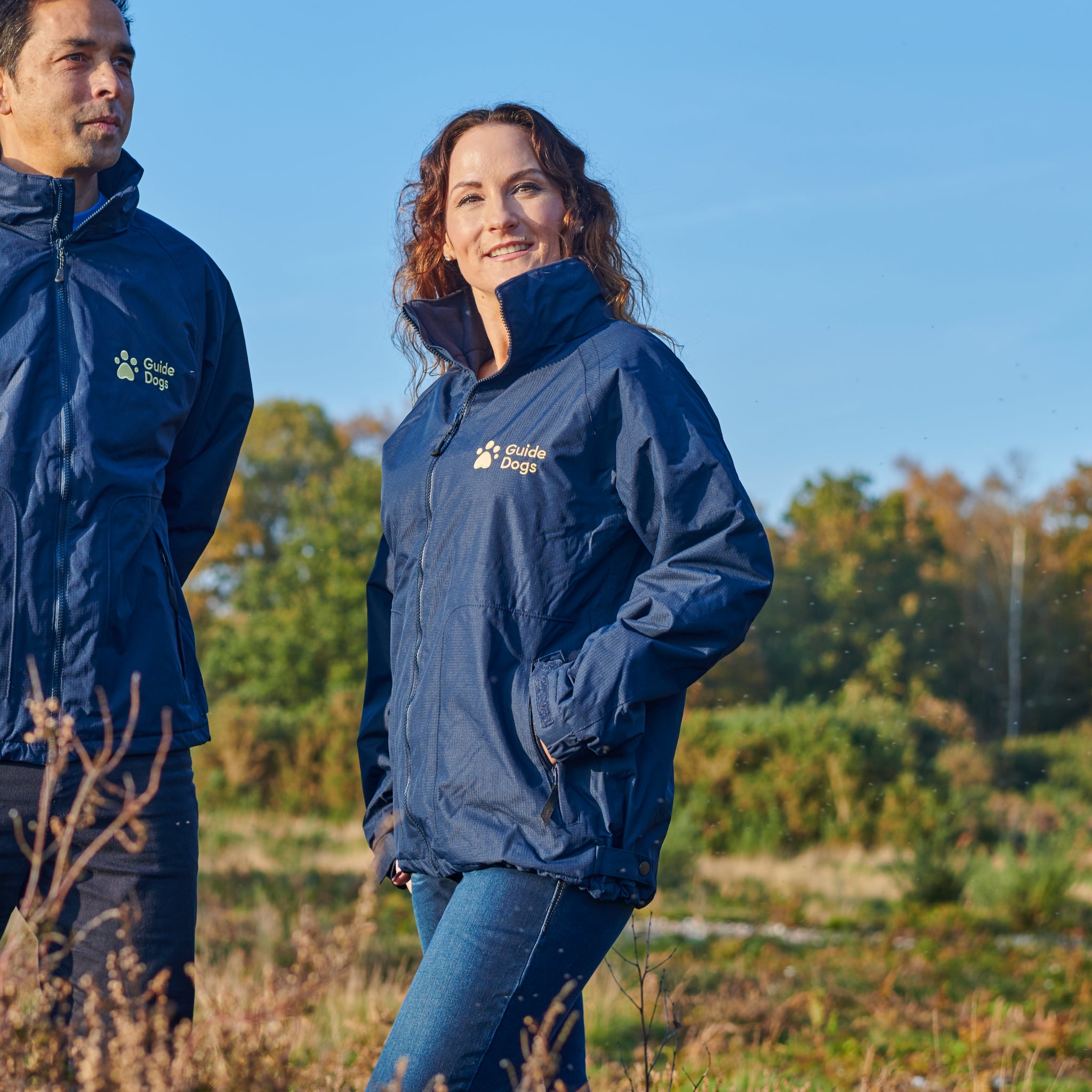 A lady with brown curly hair is wearing the blue Guide Dogs jacket and blue jeans in a field. A man next to her is also wearing the jacket.
