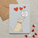 A greetings card with a Yellow Labrador with a red collar and a speech bubble saying Happy Valentines Day and three red hearts. Behind the card is a brown paper envelope on a textured background.