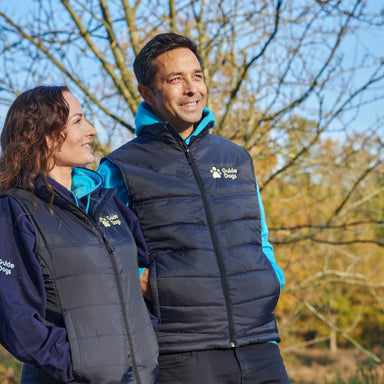 A dark haired gentleman is in front of a tree wearing a navy padded body warmer with the Guide Dogs logo in yellow on the upper left corner and a light blue hoodie. A lady next to him is wearing the same body warmer.