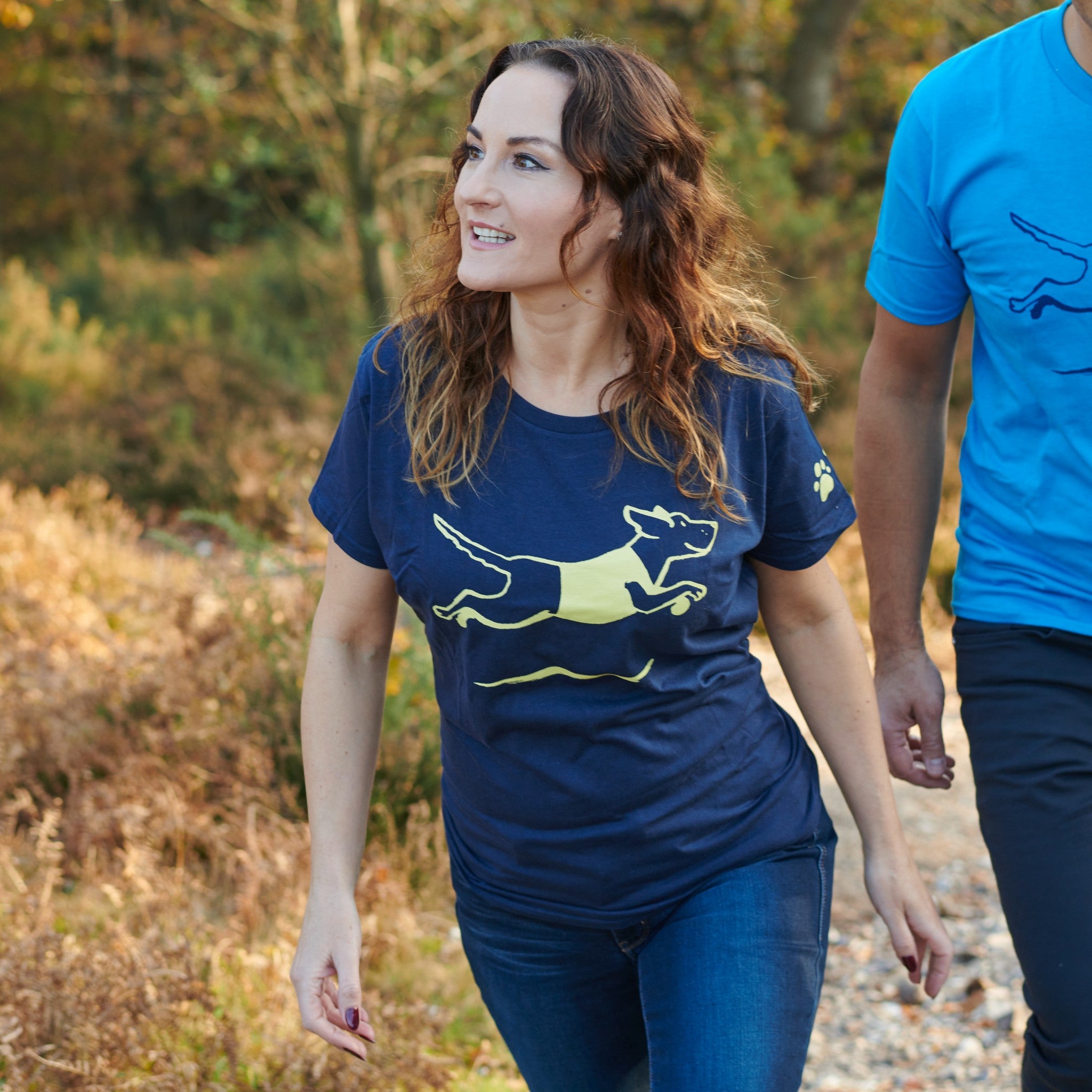 A lady with brown hair wears a navy tshirt with a leaping dog printed on the front and the Guide Dogs logo on the arm, both in yellow.